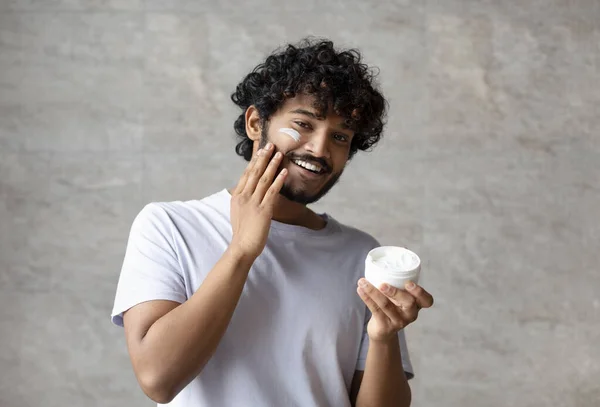 Cheerful indian man applying cream on face, holding moisturizer jar in hand, smiling to camera, standing in bathroom — Stok fotoğraf