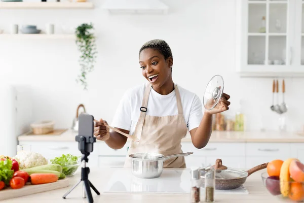 Satisfied millennial black lady with open mouth in apron prepares dinner, shows her dish, shoots video for food blog — Stock Photo, Image