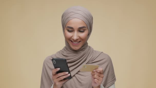 Compras por Internet. Joven mujer árabe en pañuelo para la cabeza haciendo transacción financiera en línea en el teléfono inteligente con tarjeta de crédito — Vídeos de Stock