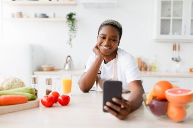 Önlüklü neşeli siyahi kadın mesaj okuyor, telefonda sebzelerle selfie çekiyor.
