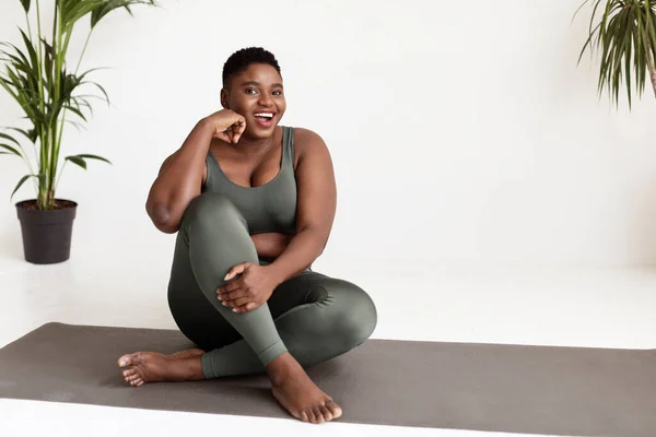 Emocionada mujer negra con sobrepeso sentada en una esterilla de yoga —  Fotos de Stock