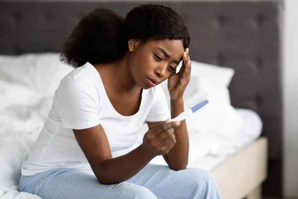 Μπερδεμένη μαύρη κυρία που κάθεται στο κρεβάτι, κρατώντας τεστ εγκυμοσύνης — Φωτογραφία Αρχείου