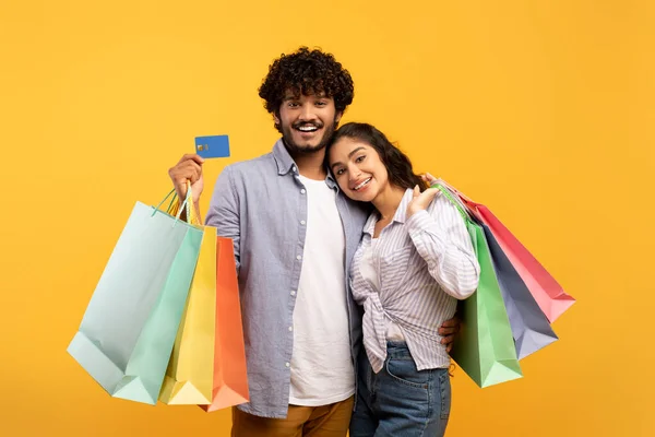 Sevgi dolu Hintli çift kredi kartı ve renkli alışveriş torbalarıyla poz veriyor, birlikte alışveriş yapıyor, sarı arka planda — Stok fotoğraf