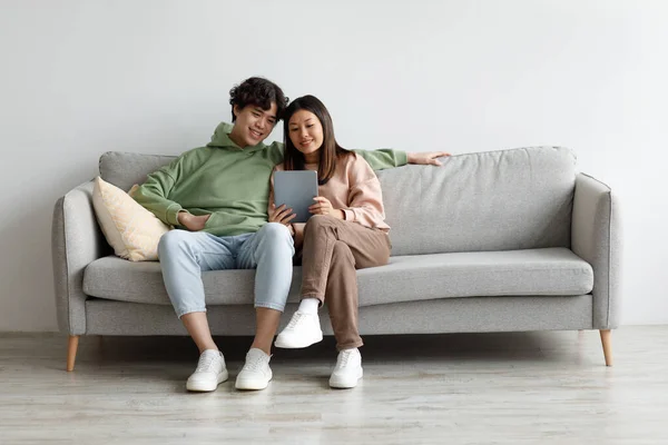 Ευτυχισμένο ασιατικό ζευγάρι κάθεται στον καναπέ με tablet, κοιτάζοντας την οθόνη αφής, χρησιμοποιώντας νεότερη εφαρμογή στο σπίτι — Φωτογραφία Αρχείου
