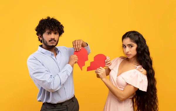 Concept de fin d'amour. Couple indien malheureux tenant des morceaux de coeur brisé sur fond jaune studio — Photo