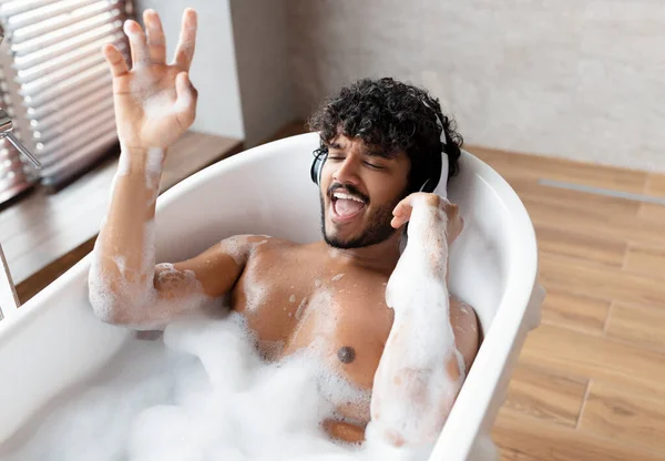 Chico indio alegre en auriculares inalámbricos disfrutando de la música y el canto mientras está acostado en el baño caliente burbujeante en casa — Foto de Stock