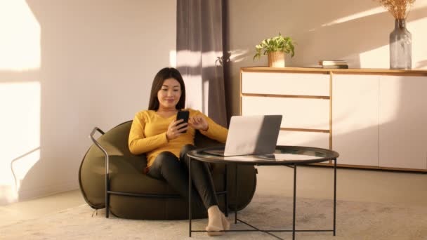 スマートフォンを使った幸せなアジア千年紀の女性が自宅で座り — ストック動画