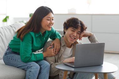 Mutlu genç Asyalı çift dizüstü bilgisayar kullanıyor, video veya film izliyor, uzaktan alışveriş yapıyor, internetten sosyal medyayı kontrol ediyor.