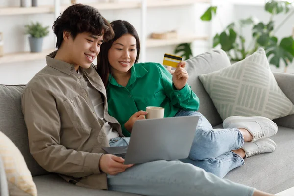 Heureux couple asiatique shopping en ligne via ordinateur portable et carte de crédit, commander des choses sur Internet, assis sur le canapé à la maison — Photo