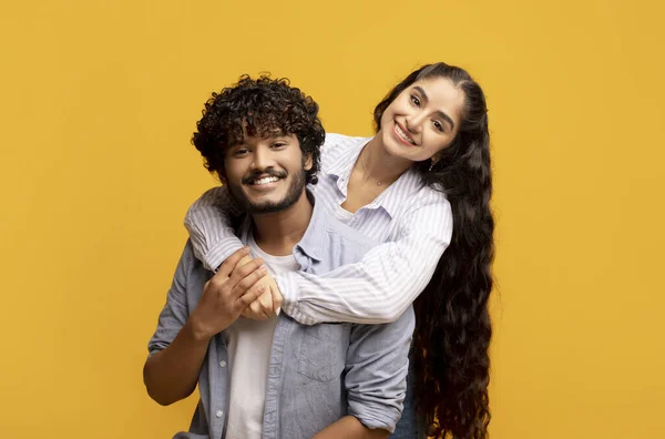 Mujer india encantadora abrazando a su marido por detrás, sonriendo y sintiéndose feliz en el fondo amarillo del estudio — Foto de Stock