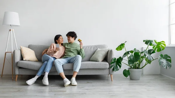 Alegre jovem casal asiático sentado no sofá com cartão de crédito e celular, compras on-line na sala de estar, espaço de cópia — Fotografia de Stock