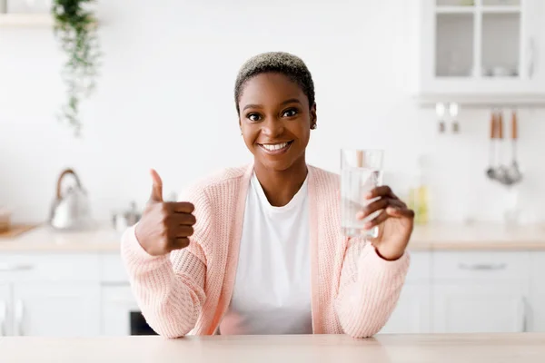 Blij lachende jonge mooie zwarte vrouw met perfecte huid houdt glas met schoon water en toont duim omhoog — Stockfoto