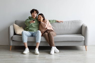 Korkak genç Asyalı çift TV 'de korku filmi izliyor, uzaktan kumanda kullanıyor, evdeki koltukta oturuyor, uzayı kopyalıyor.