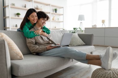 Genç Asyalı bir kadın erkek arkadaşına sarılıyor, dizüstü bilgisayar kullanıyor, evdeki kanepede internette geziniyor, boş bir alanda.