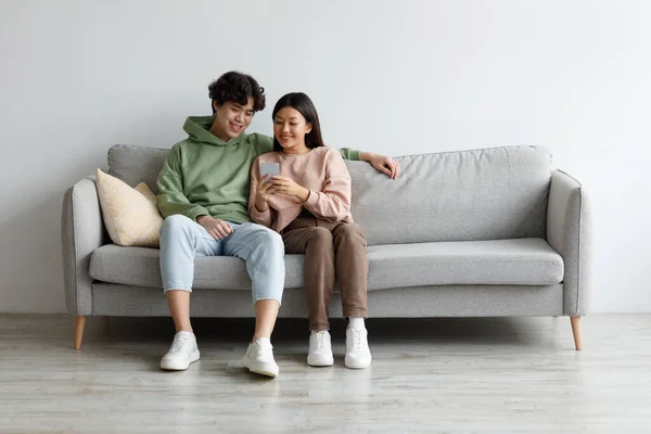 Glada unga asiatiska par som använder mobiltelefon, surfa på internet, chatta online, koppla av på soffan hemma, ledigt utrymme — Stockfoto