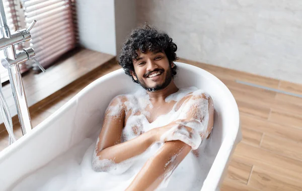 Homem indiano nu feliz tomando banho em casa, deitado na banheira branca com espuma e sorrindo para a câmera, espaço cópia — Fotografia de Stock