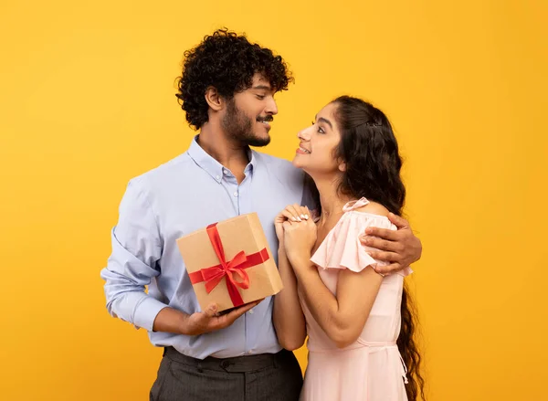 Celebration concept. Loving indian couple embracing and man holding wrapped gift box, yellow studio background — Stock Photo, Image