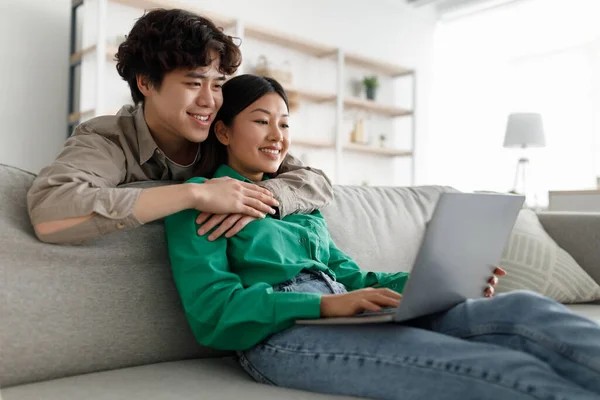 Amando jovem casal asiático usando laptop juntos, abraçando, navegando na internet, sentado no sofá em casa, espaço livre — Fotografia de Stock