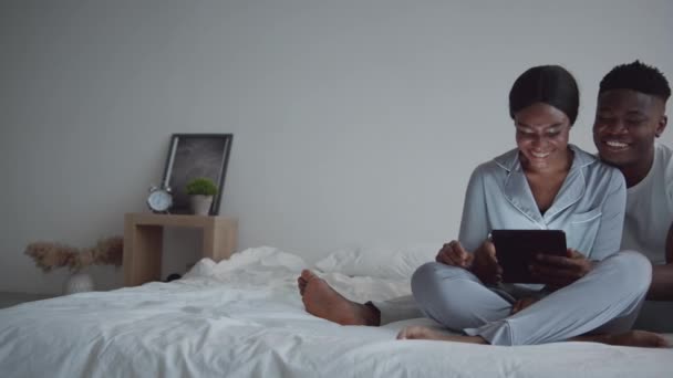 Tracking shot di interni accogliente camera da letto, amorevoli sposi afro-americani seduti sul letto e la navigazione web su tablet digitale — Video Stock