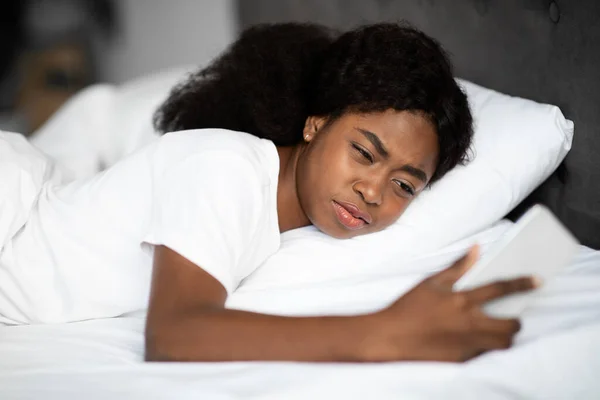 Uykucu Afrikalı Amerikalı kadın yatakta akıllı telefon kullanıyor. — Stok fotoğraf