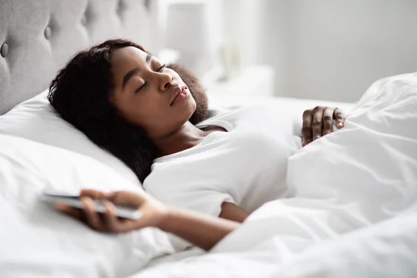 Mujer negra infeliz acostada en la cama, sosteniendo el teléfono celular —  Fotos de Stock