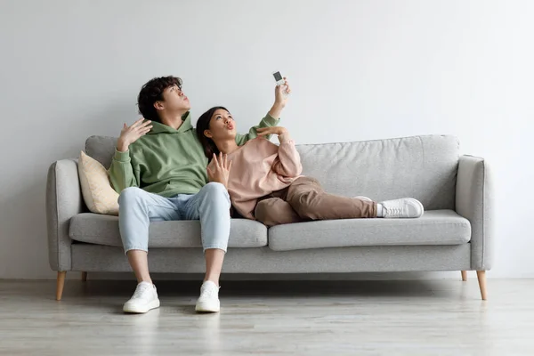 千禧年的亚洲夫妇打开空调，使用遥控装置，在家里遭受夏季炎热的折磨，模仿 — 图库照片