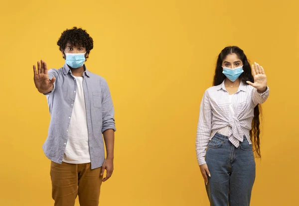 Társadalmi távolság és Covid-19 járvány. Fiatal indiai pár orvosi maszk mutató STOP gesztus, sárga háttér — Stock Fotó