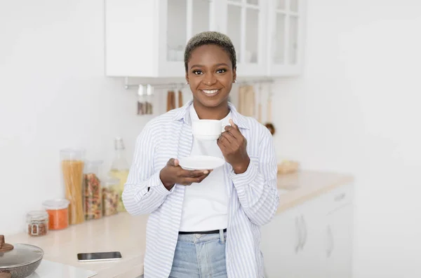 Cheerful pretty millennial black female in casual clothes drinking her favorite hot drink in white cup — Stock Photo, Image