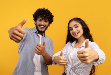 Büyük indirim. Kameraya başparmaklarını gösteren neşeli Hintli çift, sarı arka planda poz veren havalı teklif için heyecanlılar.