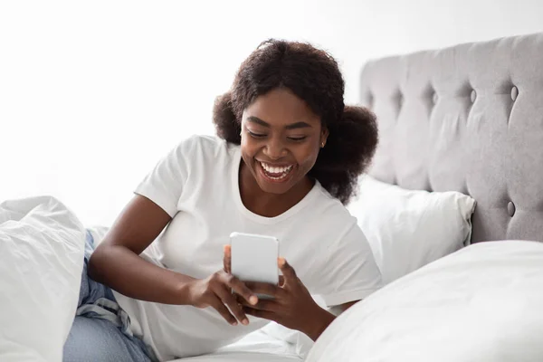 Mujer negra positiva despertando en la mañana, usando el teléfono celular —  Fotos de Stock