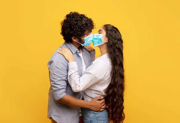 Szerelem a világjárvány idején. Fiatal indiai pár csók és ölelés, visel orvosi arc maszkok sárga háttér — Stock Fotó