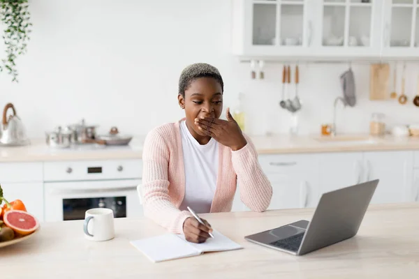 Κουρασμένη νεαρή όμορφη μαύρη γυναίκα κοιτάζει φορητό υπολογιστή κάνει σημειώσεις, χασμουρητά στο μινιμαλιστικό εσωτερικό της κουζίνας, ελεύθερο χώρο — Φωτογραφία Αρχείου