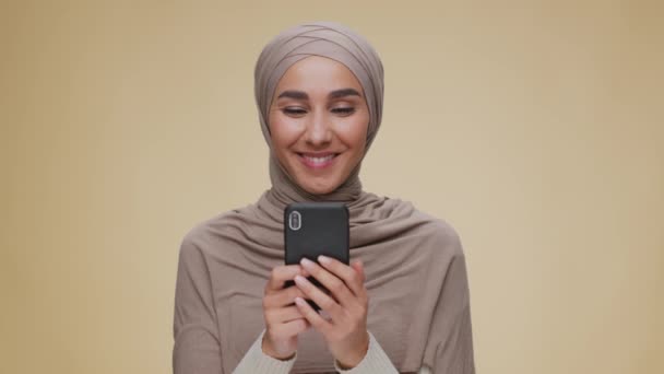 Joven mujer positiva de Oriente Medio con hijab en red o charlando en el teléfono inteligente y sonriendo, fondo beige — Vídeos de Stock