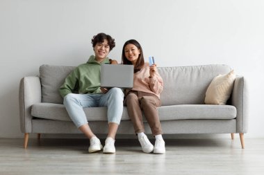 İnternetten alışveriş. Asyalı çift evdeki kanepede oturuyor, dizüstü bilgisayar ve kredi kartı kullanıyorlar, web mağazasından mal alıyorlar.
