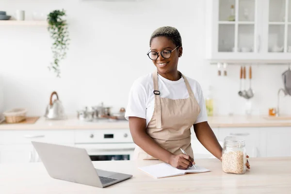 Cheerful millennial pretty black female in glasses, apron writes recipe looks pc in minimalist kitchen interior — Stock Photo, Image