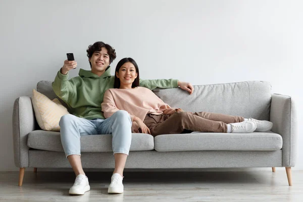 Jeune couple asiatique se détendre sur le canapé, regarder la télévision, changer de chaînes avec télécommande, profiter du film à la maison — Photo