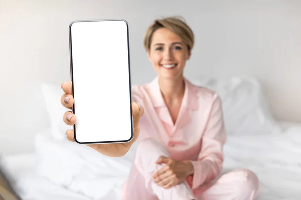Bella donna seduta a letto e mostrando grande smartphone vuoto alla fotocamera — Foto Stock