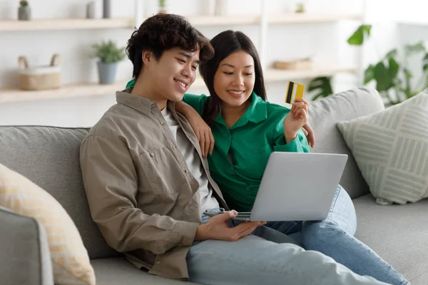 Позитивна азіатська сім'я займається онлайн-магазинами з кредитною карткою і ноутбуком, сидячи на дивані і обіймаючись вдома. — стокове фото