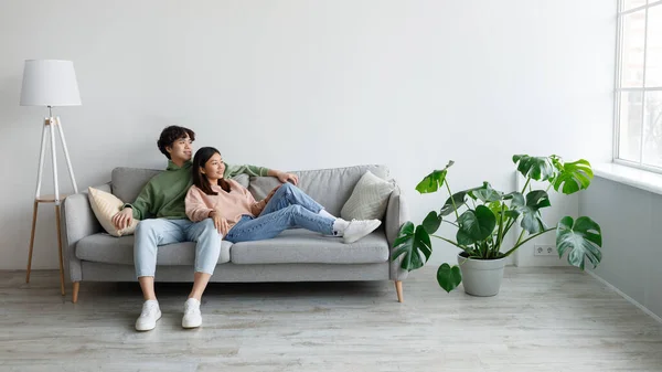 Positivo jovem casal asiático relaxante no sofá, abraçando e olhando para fora janela na sala de estar, espaço livre — Fotografia de Stock