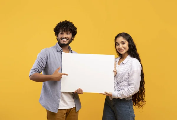 Lugar para o seu anúncio. Retrato de casal indiano segurando placa de cartaz vazia em branco, apontando para ele sobre fundo amarelo — Fotografia de Stock