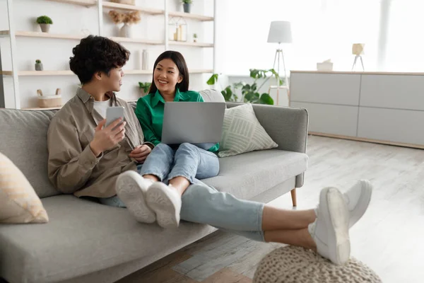 Asiatisches Paar benutzt Laptop und Smartphone zu Hause, kuschelt auf der Couch, schaut Videos, surft im Internet, shoppt online — Stockfoto