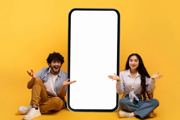 Förvånad indian par sitter nära stora smartphone med tom skärm över gul studio bakgrund, mockup — Stockfoto