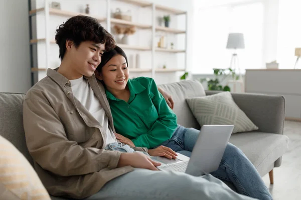 Conceito de passatempos de fim de semana. Casado jovem casal asiático usando laptop em casa, relaxando no sofá juntos, espaço de cópia — Fotografia de Stock