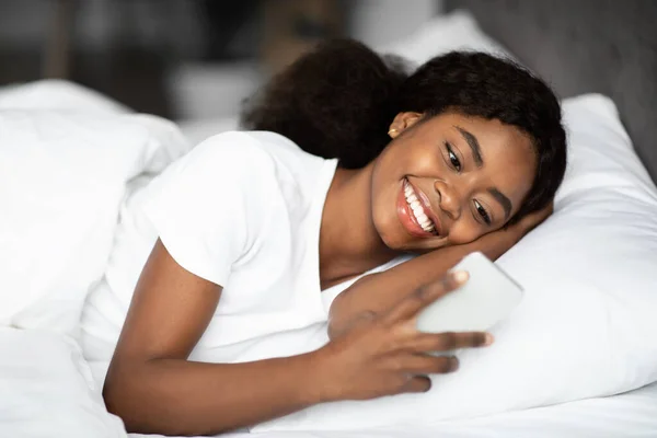 Feliz joven negra acostada en la cama con teléfono inteligente —  Fotos de Stock