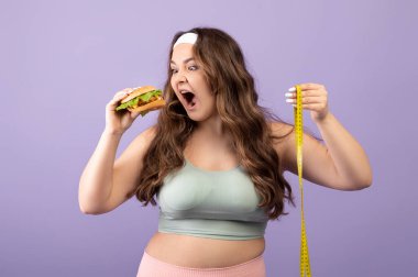 Komik, aç, beyaz, spor giyimli, spor kıyafetli genç bir kadın mezeci burgeri yiyor.