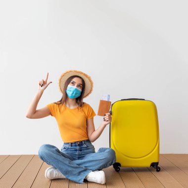 Turist teklifi. Koruyucu maskeli kadın ışık duvarının üzerinde boş bir alanı işaret ediyor, valizin yanında oturuyor.