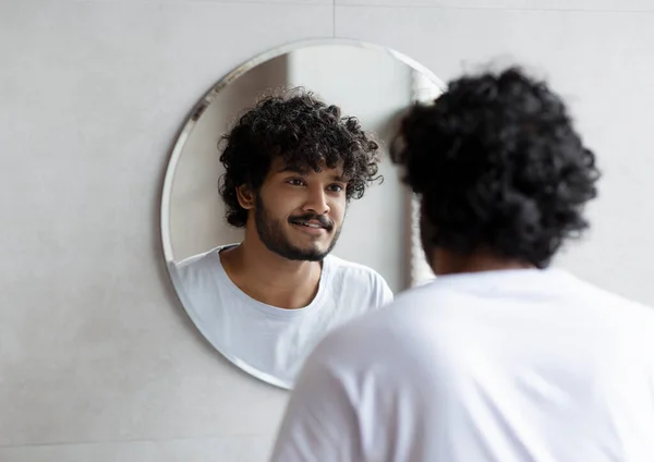 Tipo indio guapo mirando su reflejo en el espejo, de pie en el baño moderno en casa —  Fotos de Stock