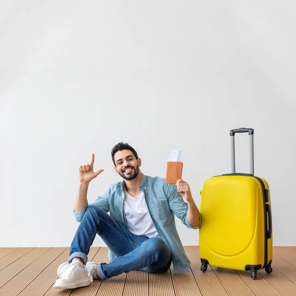 Happy Araab man met vliegtickets, paspoort en koffer poserend over de lichtmuur en wijzend naar kopieerruimte — Stockfoto