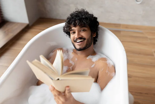 Boldog indiai férfi fekszik habos fürdő és olvasókönyv pihentető légkörben a fürdőszoba belsejében, szabad hely — Stock Fotó