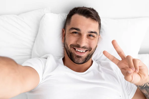 Glimlachende duizendjarige aantrekkelijke mannelijke blogger met baard wakker op wit bed toont vrede teken en maakt selfie — Stockfoto